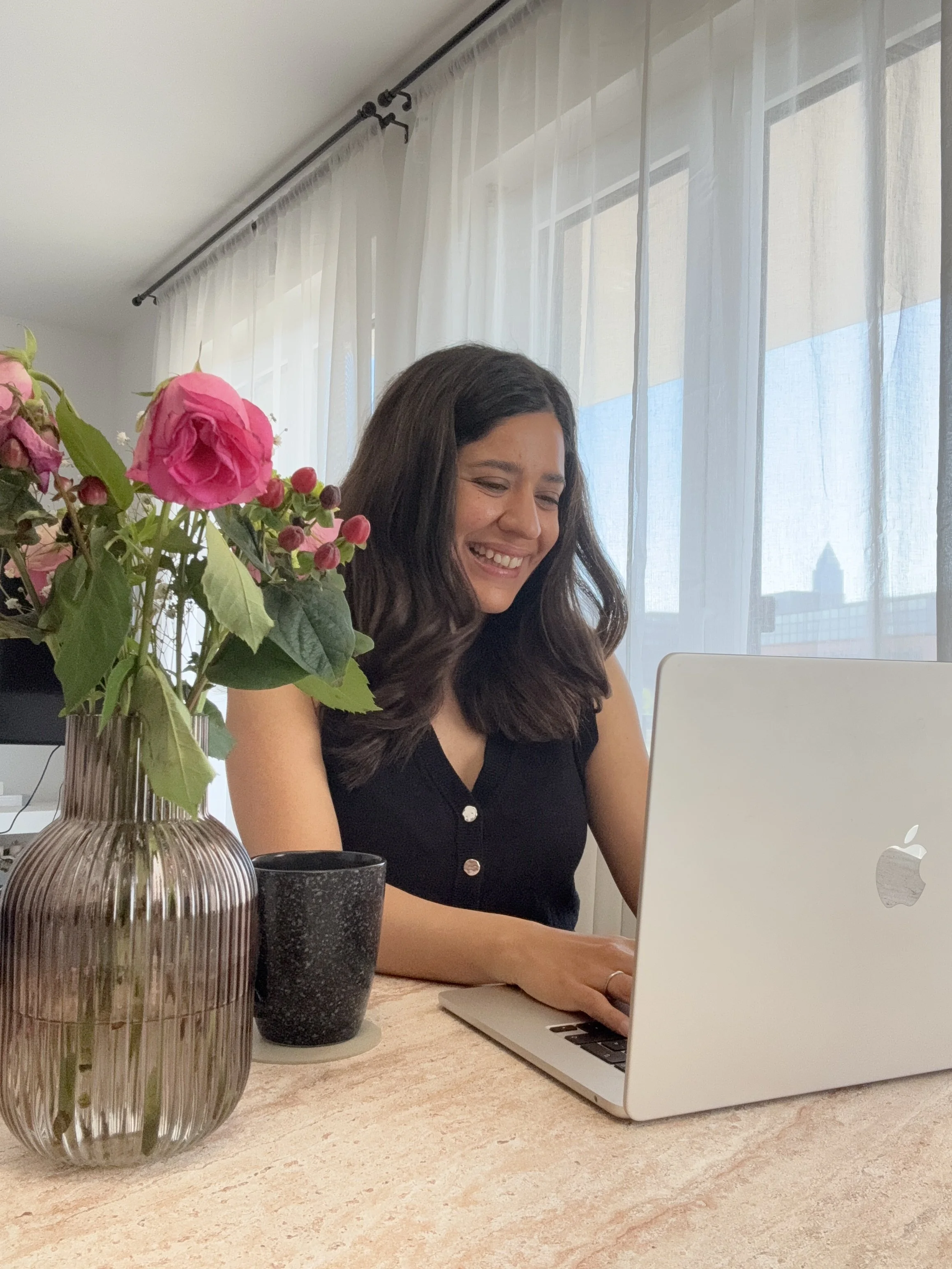 A woman with dark, curly hair smiling while working on a laptop on a desk. There are pink and red flowers in a brown vase and two black cups on the desk. Large windows with sheer white curtains are in the background.