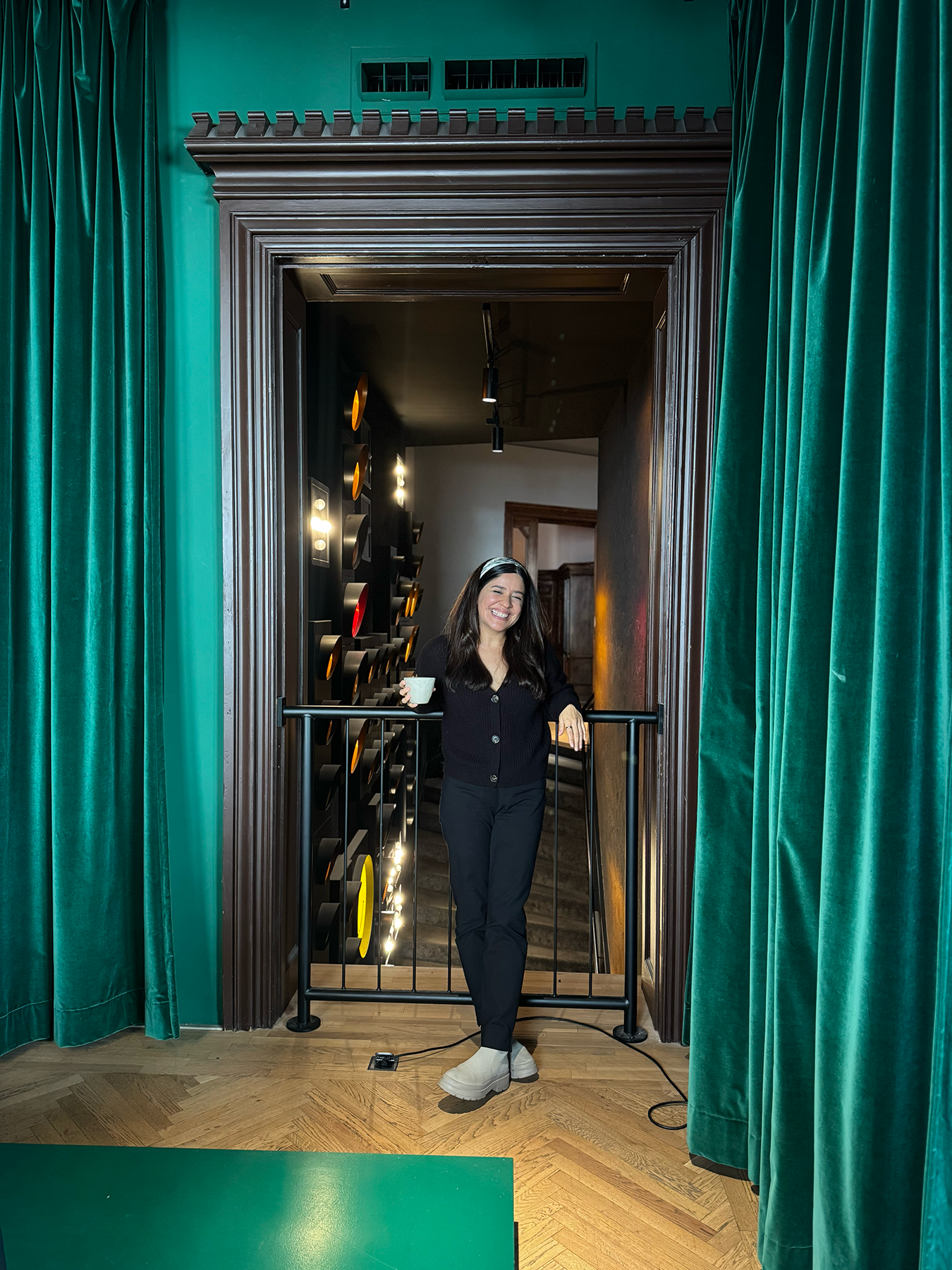 A woman with long dark hair, wearing a black sweater, black pants, and beige boots, standing in a hallway with green curtains on either side. She is smiling and holding a white cup in her right hand, leaning on a black railing. Behind her is a wall with circular, colorful decorations and lights, and a staircase leading down.