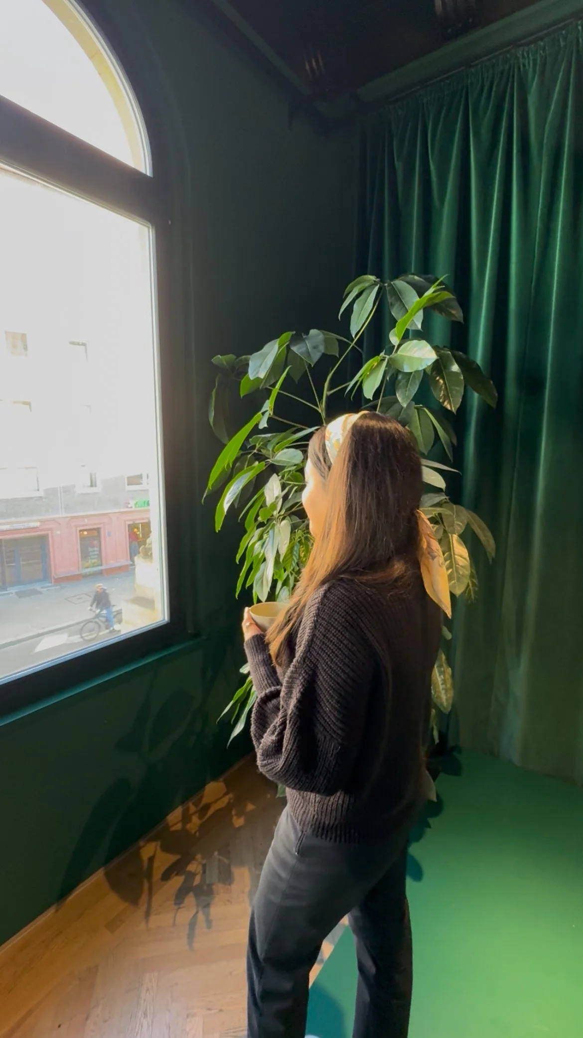 A woman with long dark hair wearing a black sweater and gray pants stands near a window holding a cup, with large green curtains and a potted plant behind her.