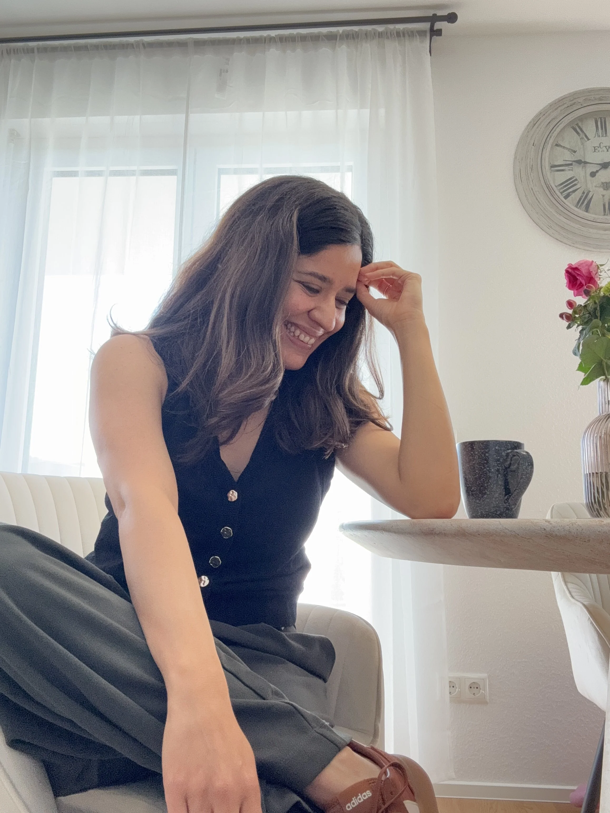 A woman with long brown hair, smiling and looking down, seated at a table in a bright room with large windows and white curtains. There is a pink flower in a vase and a dark mug on the table.
