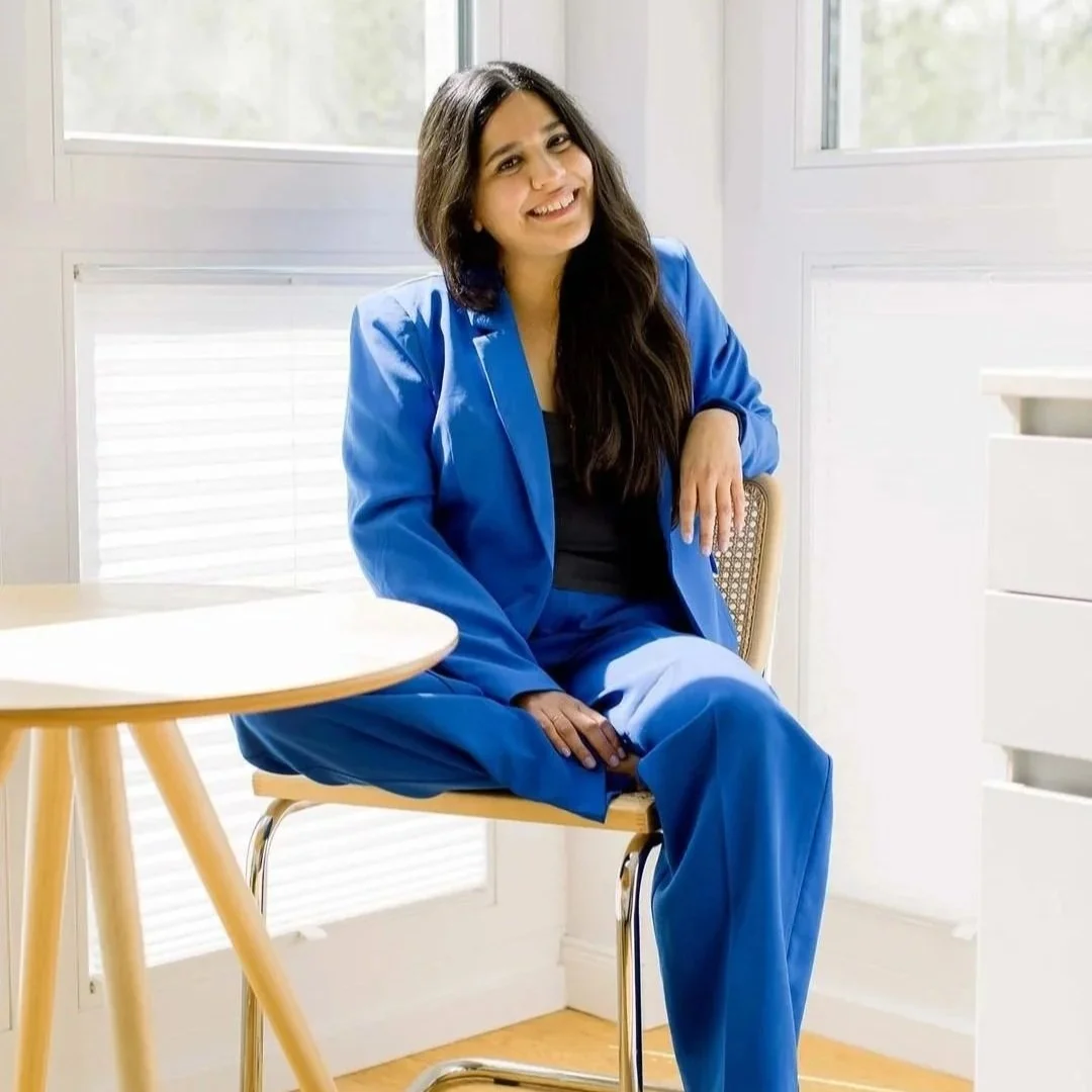 Woman with long dark hair wearing a blue suit, smiling, sitting on a chair near a window in a bright room.