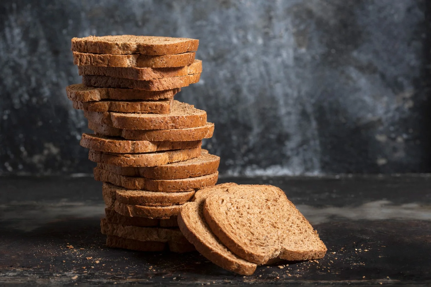 Creative studio photography of a tall stack of whole wheat bread slices on a dark background, highlighting texture and bakery craft by Stavros Kostakis.