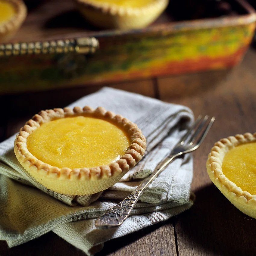Professional pastry photography by Stavros Kostakis of artisanal lemon curd tartlets on a rustic linen napkin with a vintage fork, highlighting the smooth texture and golden crust.