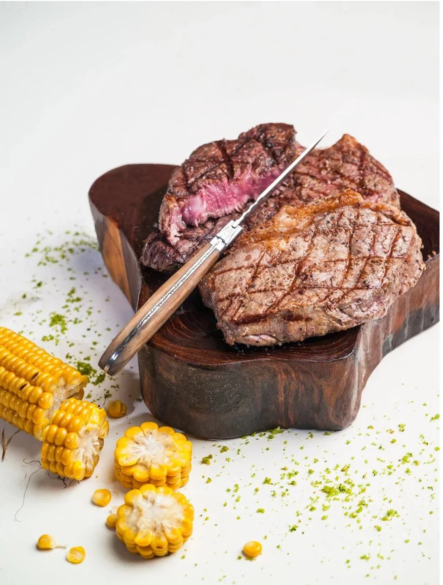 Top-down high-end commercial food photograph of a grilled steak on a wooden board with charred corn. Clean style from Stavros Kostakis' archive of commercial work in Holland. High focus on texture and composition.