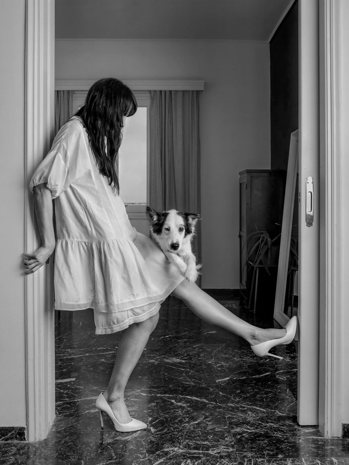 andid black and white character portrait of Antonia Katsaros in a white dress, playfully posing in a doorway with her dog. The image captures a sense of motion and genuine joy in a minimalist home setting. A lifestyle fine art study by Stavros Kostak