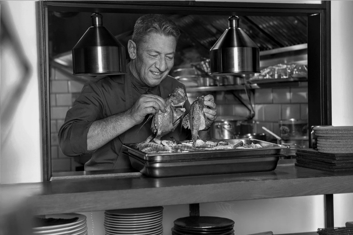 Dynamic black and white environmental portrait of Chef Alain Parodi working in the kitchen of Veritable. This character study by Stavros Kostakis captures the energy and focus of a professional chef in action, using dramatic lighting to emphasize the