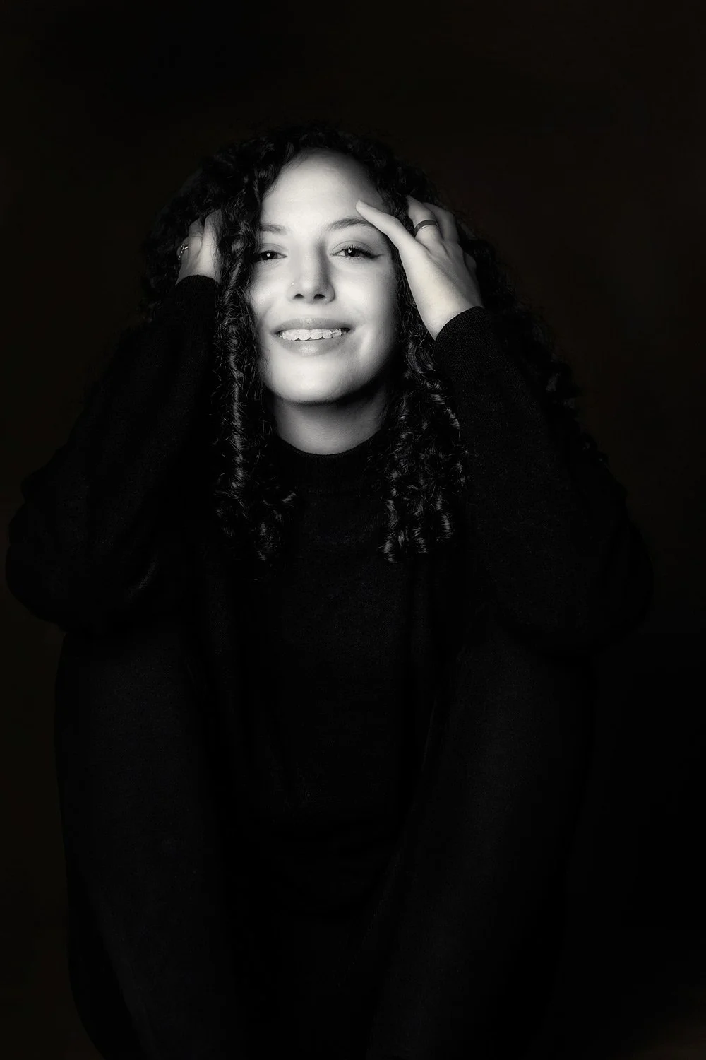 Elegant black and white studio portrait of a young woman with curly hair, featuring dramatic low-key lighting. This fine art character study by Stavros Kostakis emphasizes natural beauty, expressive emotion, and the artistic interplay of light and sh