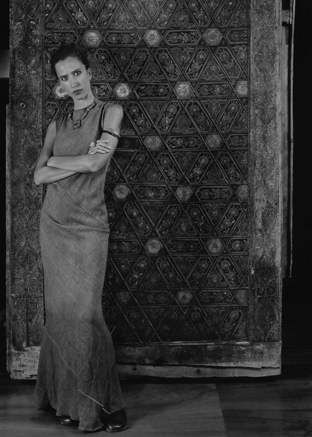 Black and white portrait of Marina Koriolanou-Lykourezou standing against an ornate carved wooden door, with a strong expression and crossed arms. Photography by Stavros Kostakis.