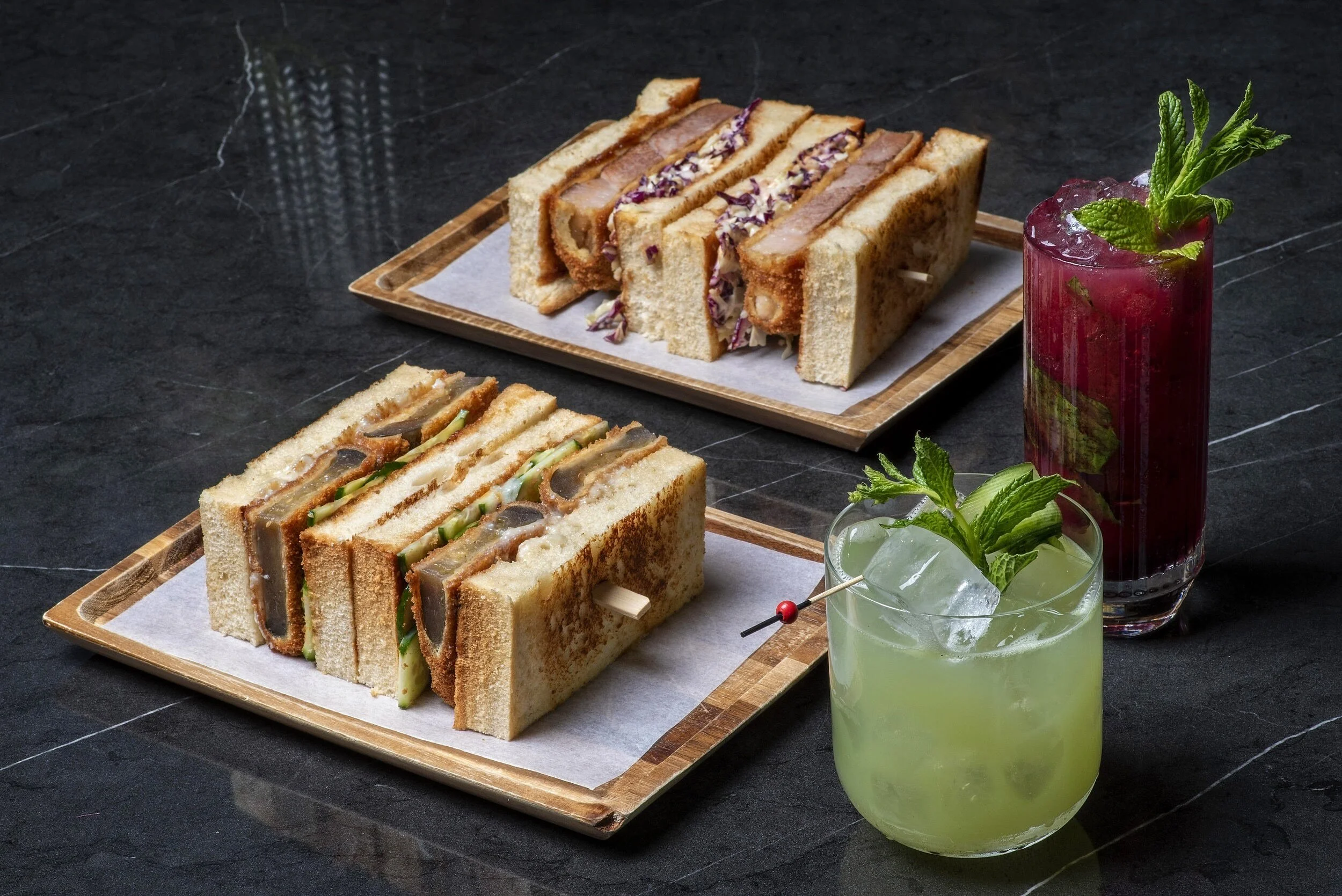 A dynamic overhead shot on black marble featuring two fusion Katsu Sandos on wooden trays, two pulled pork Bao Buns, a red berry cocktail, and a green mint cocktail. Lifestyle gastronomy photography at Royal Monkey Kifisia by Stavros Kostakis Athens.