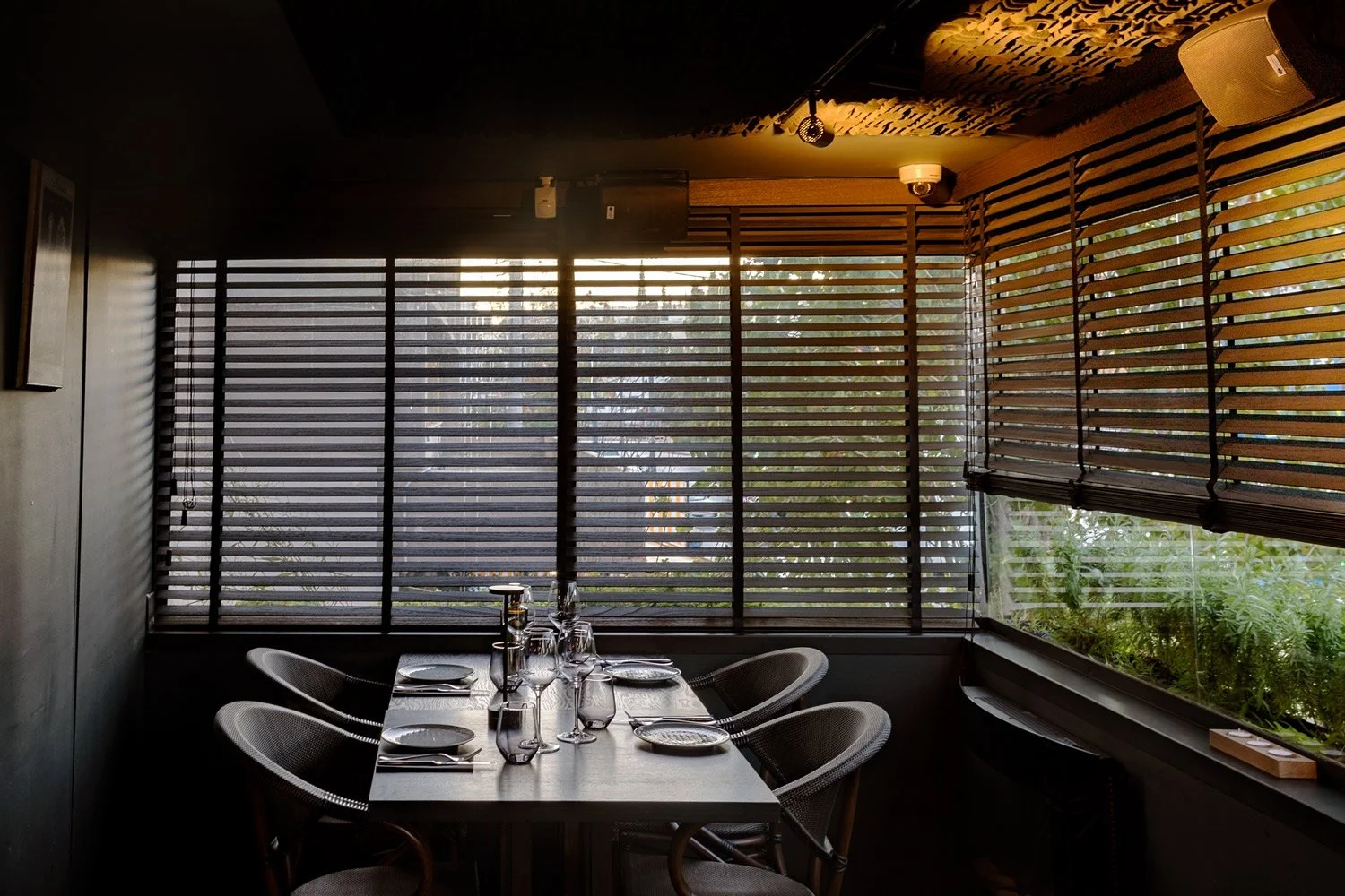 Atmospheric view of a set dining table at Veritable restaurant, surrounded by wooden Venetian blinds creating dramatic light and shadow patterns. Cinematic interior photography in Psychiko by Stavros Kostakis.
