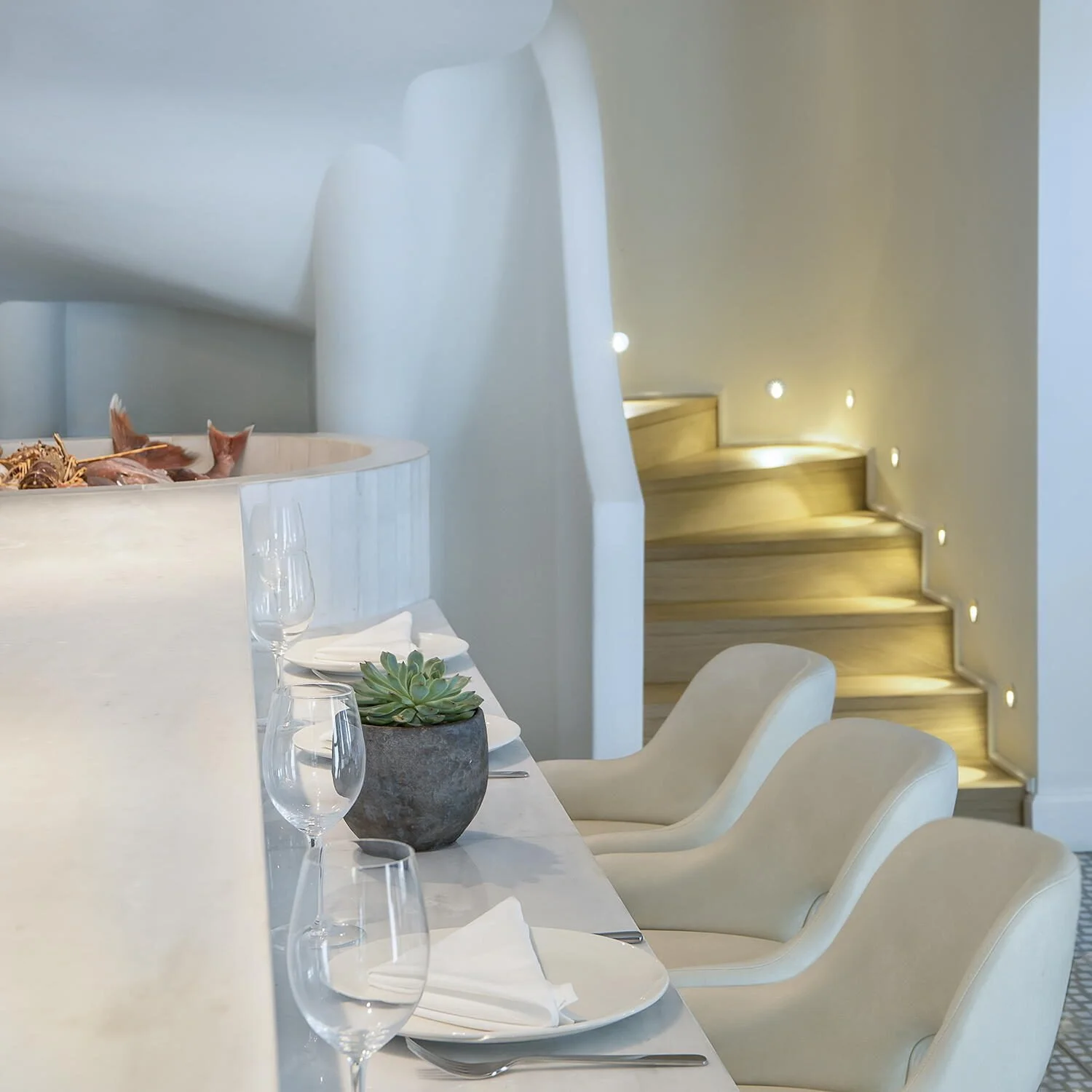 
A close-up architectural detail of the staircase area in Papaioannou restaurant, featuring warm, recessed step lighting. The shot emphasizes the soft, sculpted curves of the white walls and the clean, rhythmic perspective of the stairs, captured wit