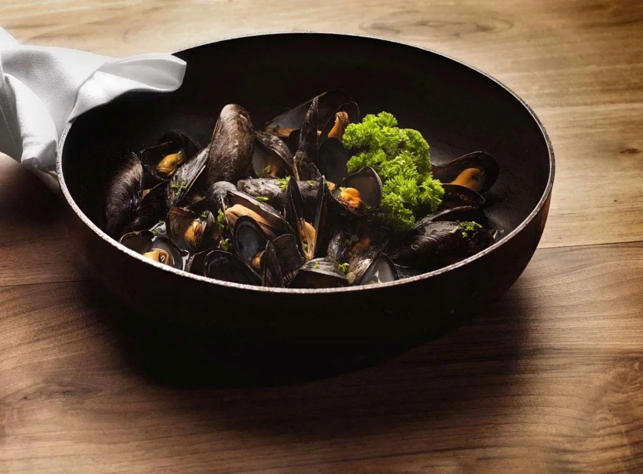 A dramatic, dark and moody culinary capture of fresh mussels served in a traditional iron skillet at Irodion Restaurant, Capelle aan den IJssel. This shot by Stavros Kostakis highlights the rich textures of the seafood and the rustic elegance of the 