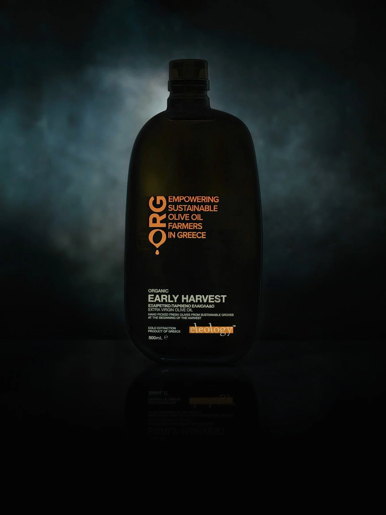 Luxury olive oil bottle photography by Stavros Kostakis for Eleology, featuring dramatic lighting, orange typography, and a perfect mirror reflection.