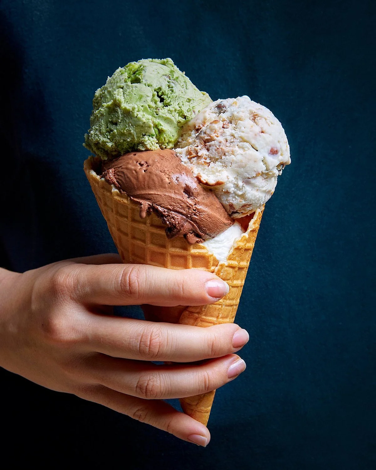Handheld artisan ice cream cone with pistachio, chocolate, and cream scoops against a dark background, professional studio lighting by Stavros Kostakis.