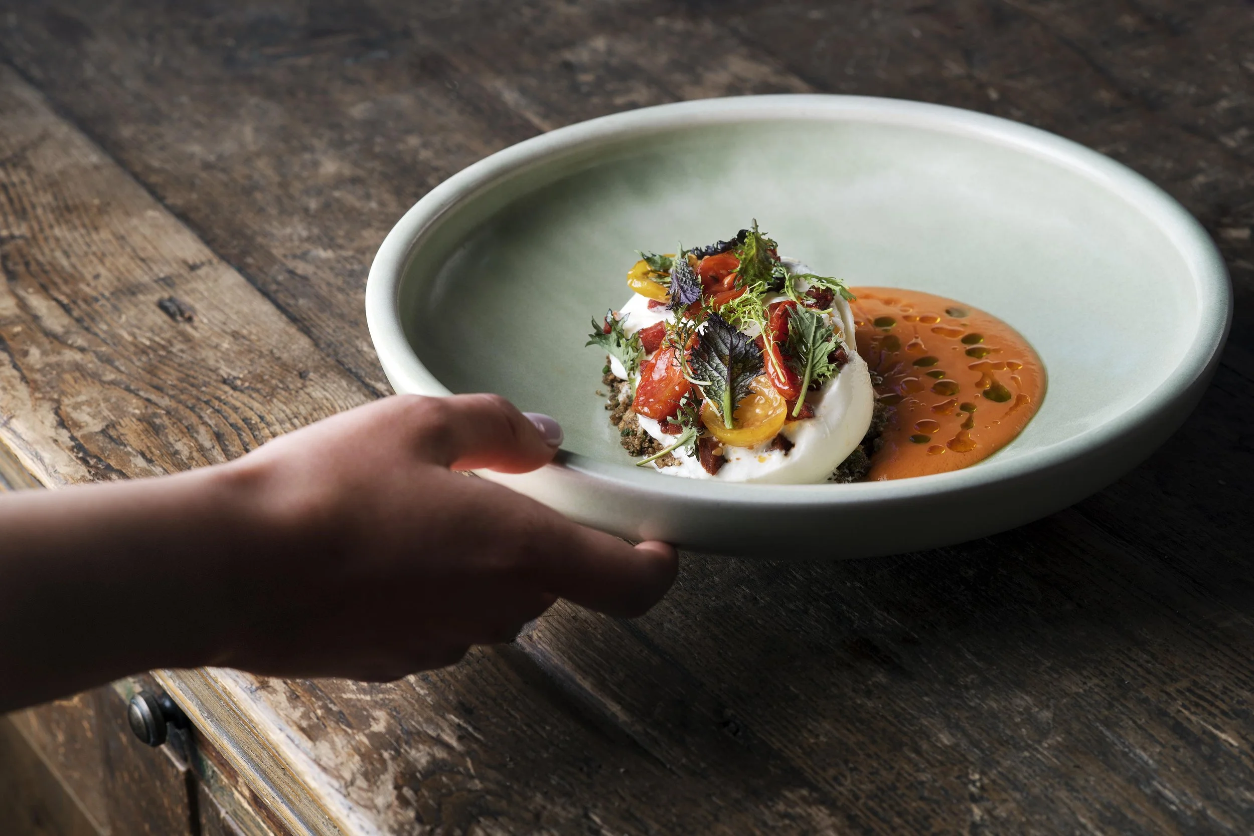 High-end lifestyle editorial food photography. A human hand gently touches a gourmet plate at Hide & Seek restaurant, creating a sense of movement and professional service. Captured by Stavros Kostakis.