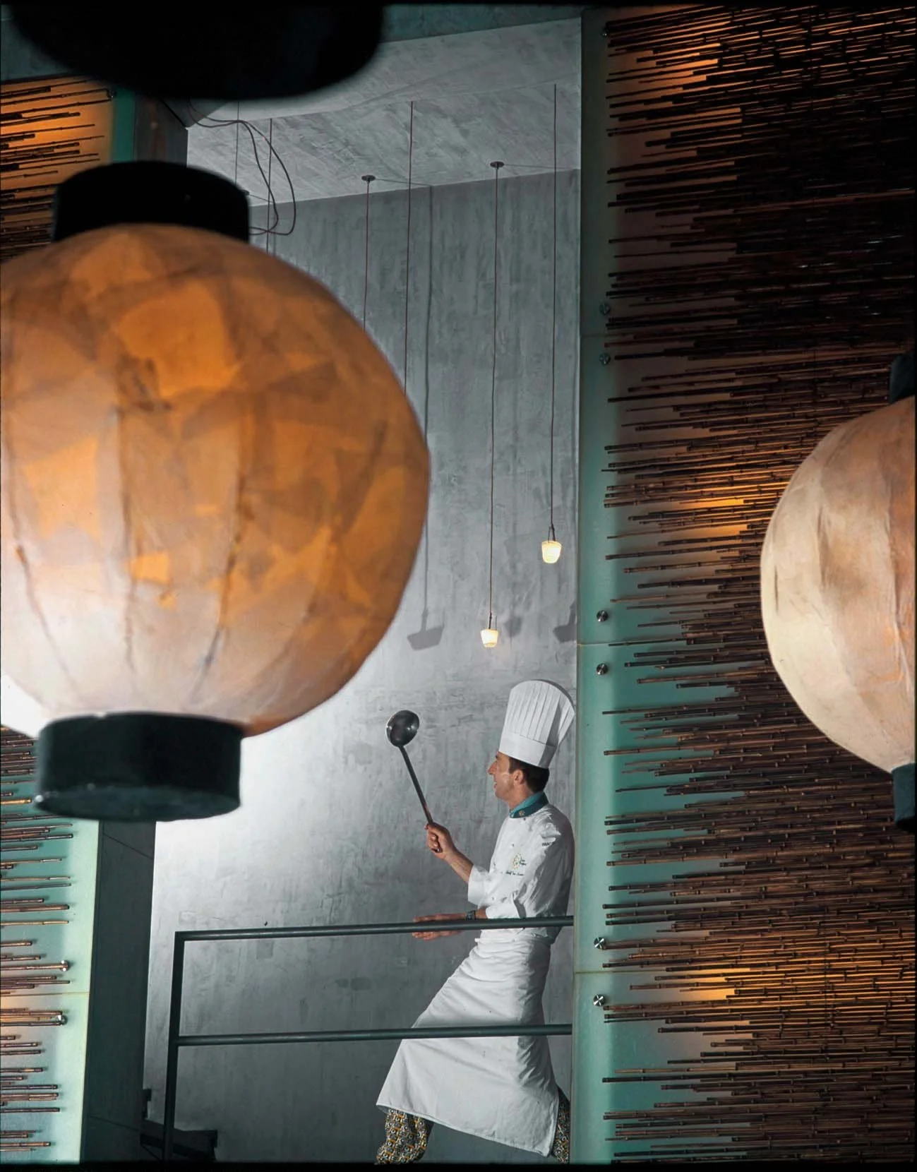 Cinematic environmental portrait of Chef Rudolf van Nunen in a sophisticated restaurant setting. The image features large warm-toned lanterns in the foreground, framing the chef as he works with precision. High-end culinary photography by Stavros Kos