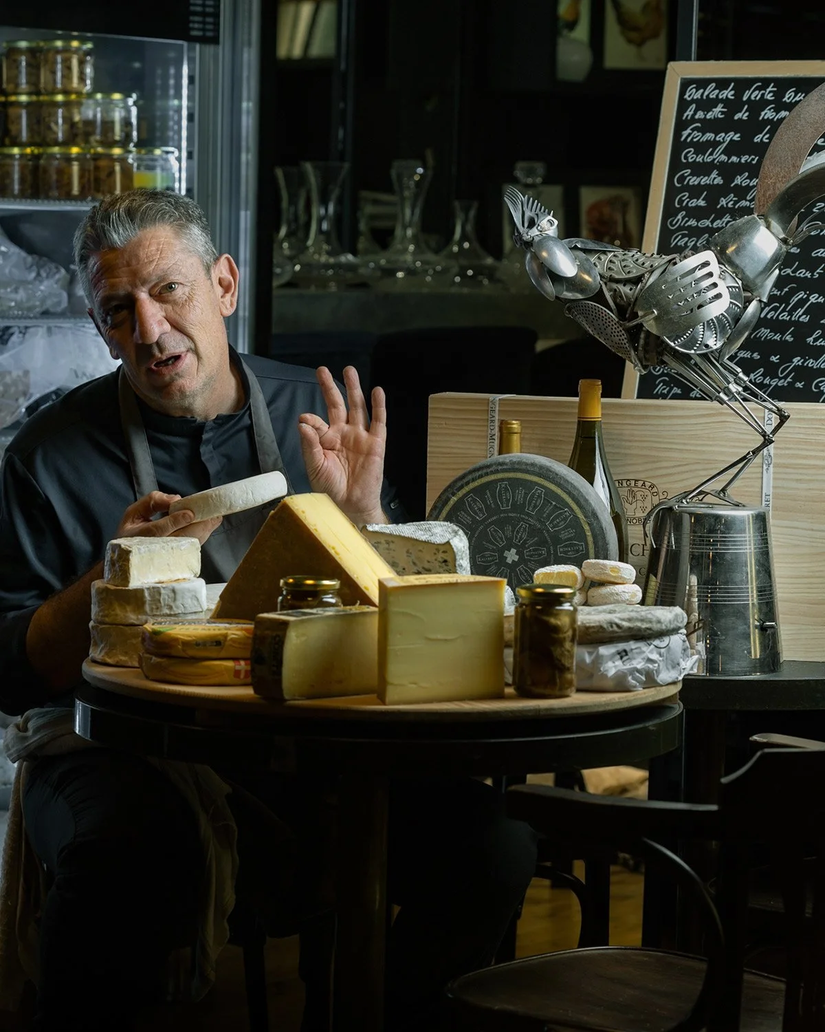 Authentic color portrait of Chef Alain Parodi in his restaurant, Veritable. The chef is captured in a candid, expressive moment behind a table filled with artisanal cheeses. High-end gastronomy photography by Stavros Kostakis focusing on the passion 
