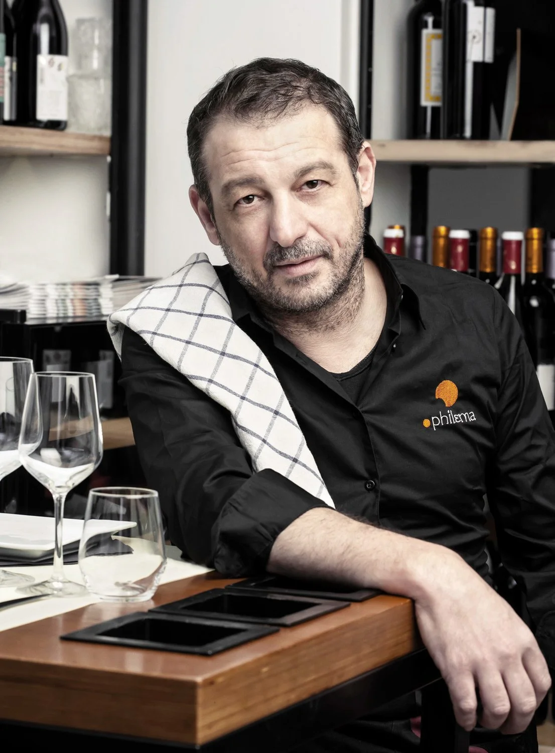 Close-up portrait of Chef Manos Makrygiannakis of Philema restaurant in Brussels. He is leaning on a wooden table with wine bottles in the background. Photography by Stavros Kostakis.