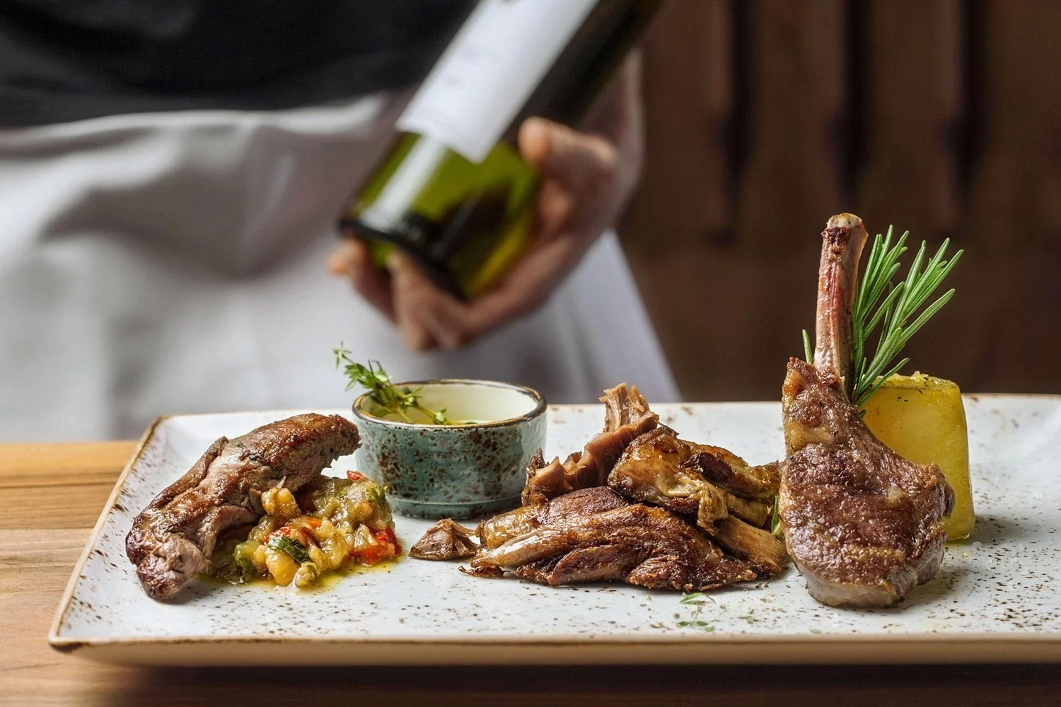 Fine Greek dining experience at Irodion Capelle, Netherlands. A close-up of a lamb cutlet dish on a wooden table, with a waiter holding a wine bottle in the background, captured by Stavros Kostakis.