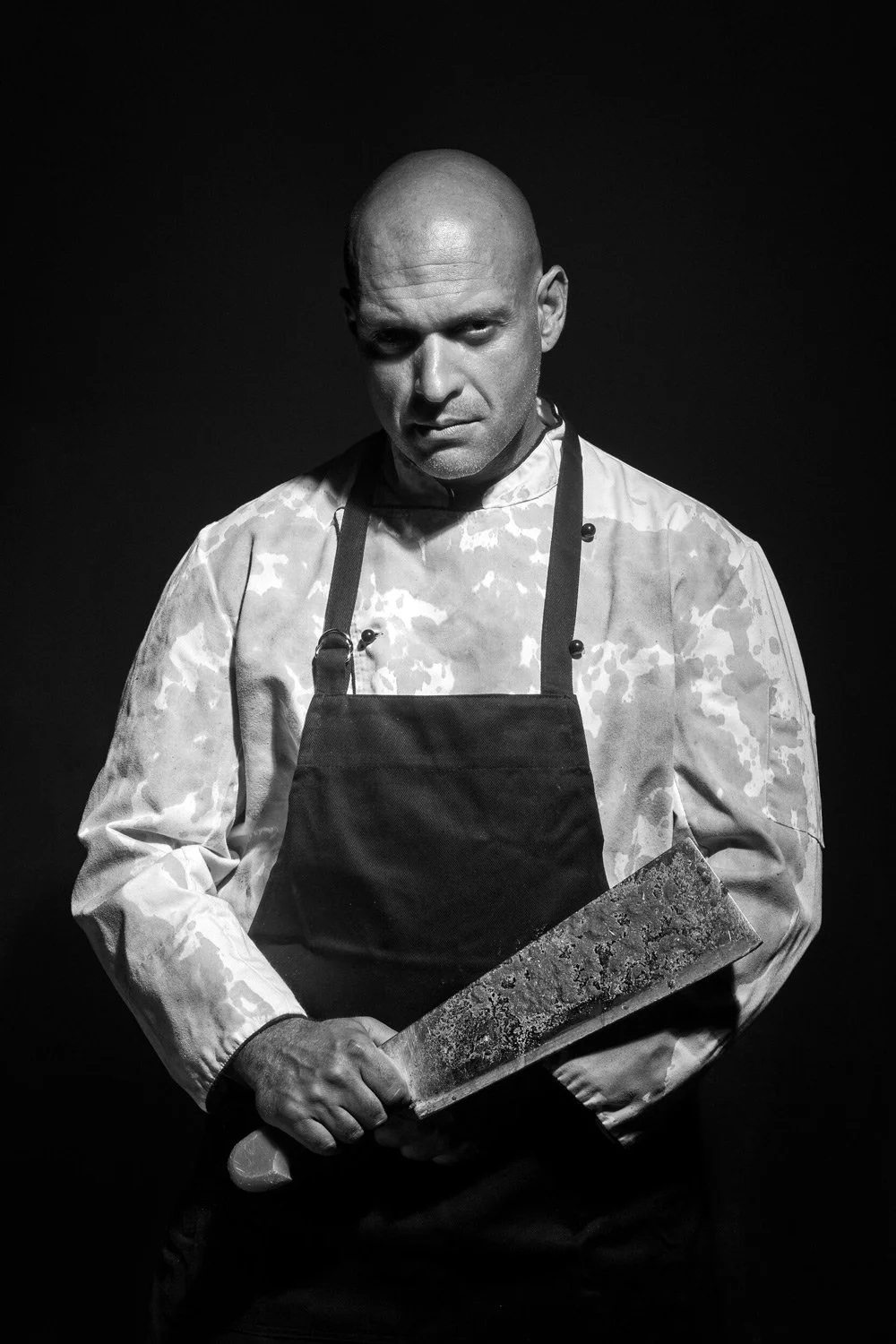 Stratos Drakoulis | Intense black and white character portrait of Stratos Drakoulis holding a large, textured meat cleaver. The lighting is dramatic and high-contrast, highlighting his stern expression and the sharp details of the blade. A powerful f