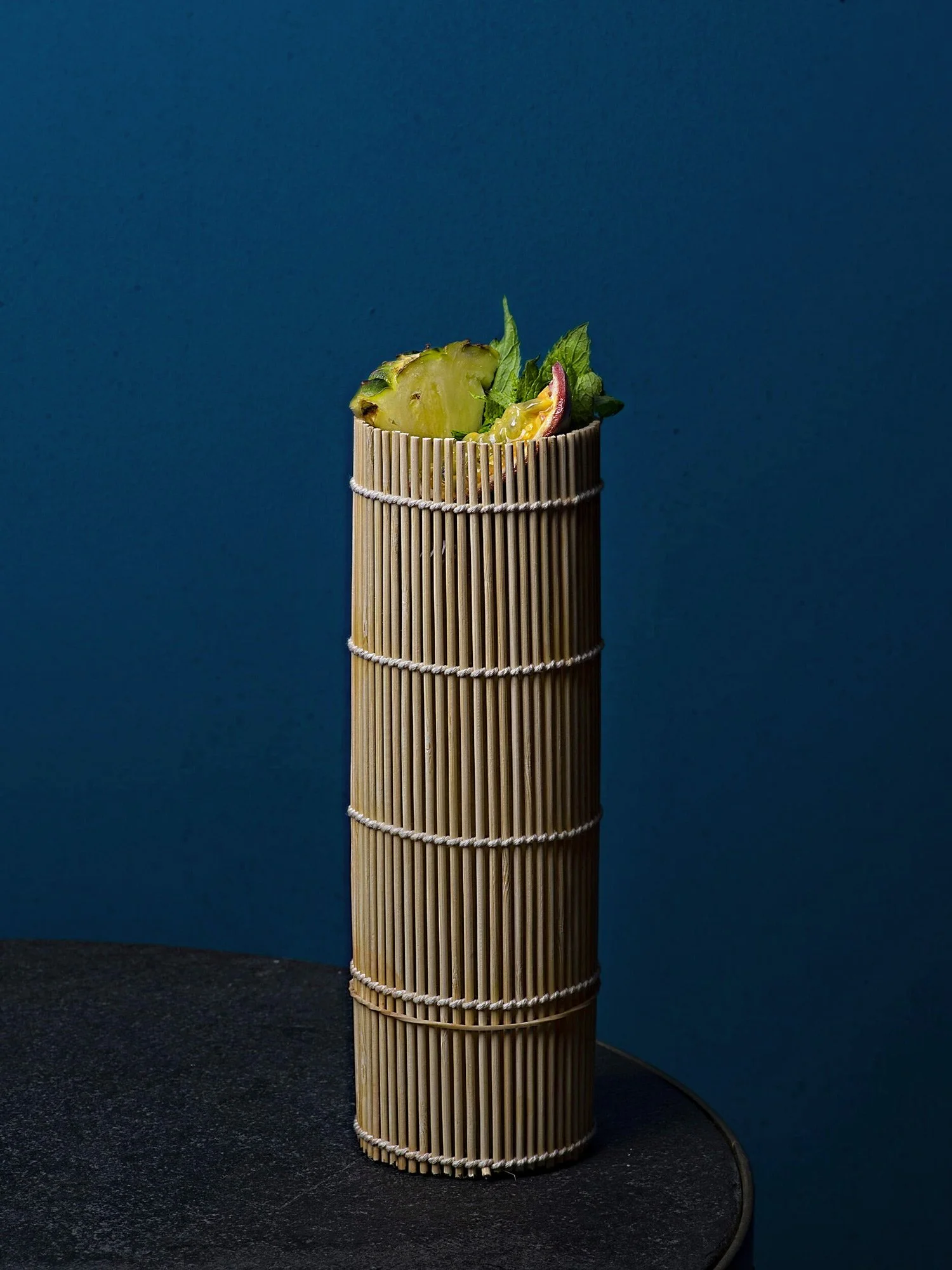 An exotic tiki-style cocktail served in a unique bamboo-textured tall glass, garnished with fresh pineapple and mint against a deep blue background at Royal Monkey Kifisia. Captured by Stavros Kostakis.