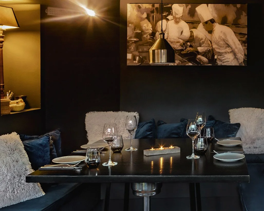Elegant interior of Veritable restaurant in Psychiko, featuring a set dining table with candles and wine glasses. The dark, moody atmosphere is highlighted by a black-and-white culinary photograph on the wall, captured by Stavros Kostakis.