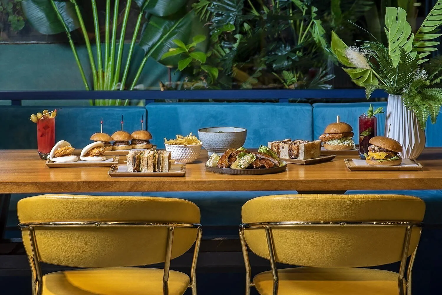 An atmospheric wide shot of Royal Monkey Kifisia's interior featuring mustard yellow velvet chairs, teal blue booths, and a lush botanical background. The wooden table is set with a fusion feast of burgers, sliders, and cocktails. Gastronomy and inte