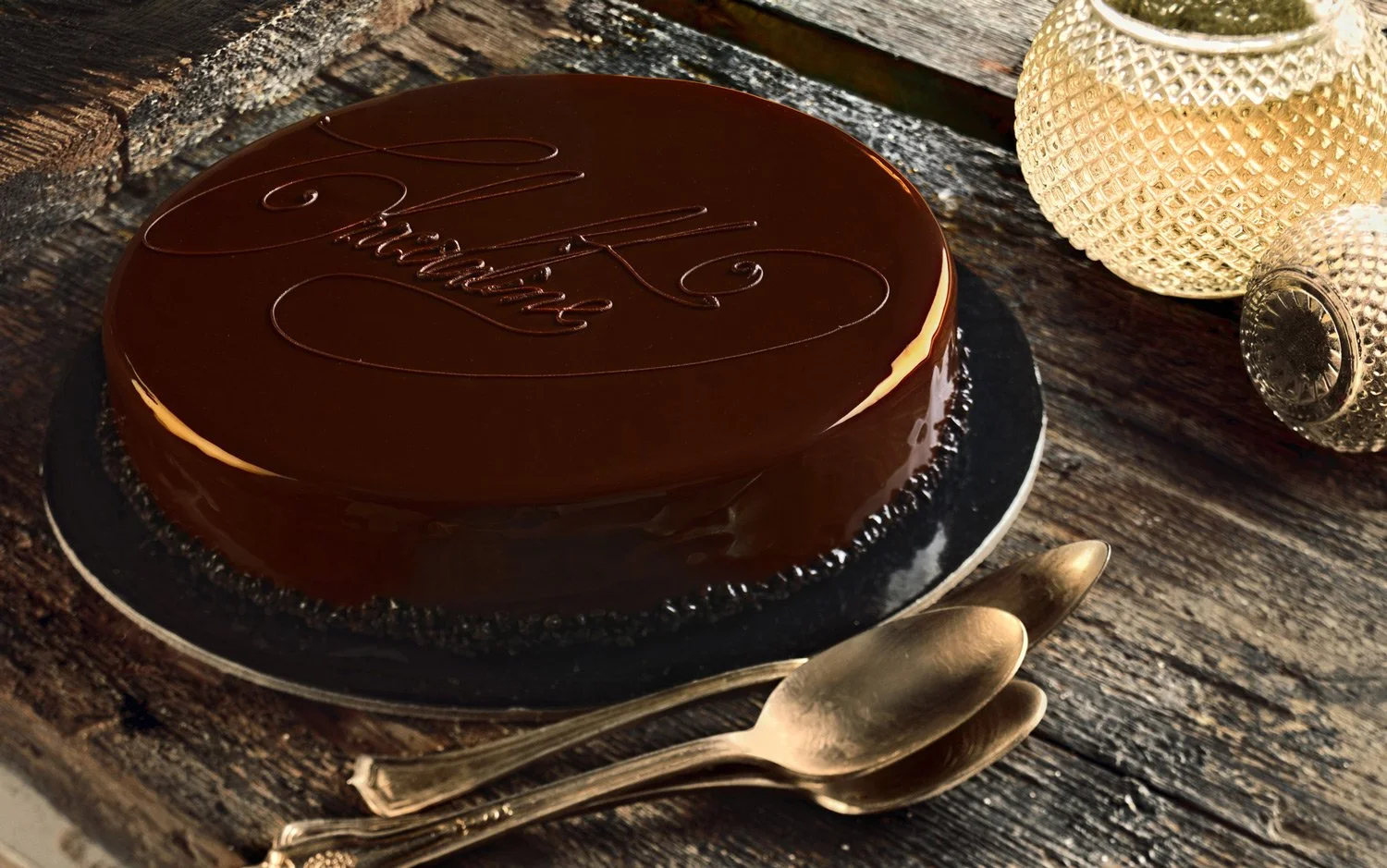 Professional food photography of a glossy chocolate glazed cake with golden spoons on a wooden surface for Papaspirou Bakery, captured by Stavros Kostakis.