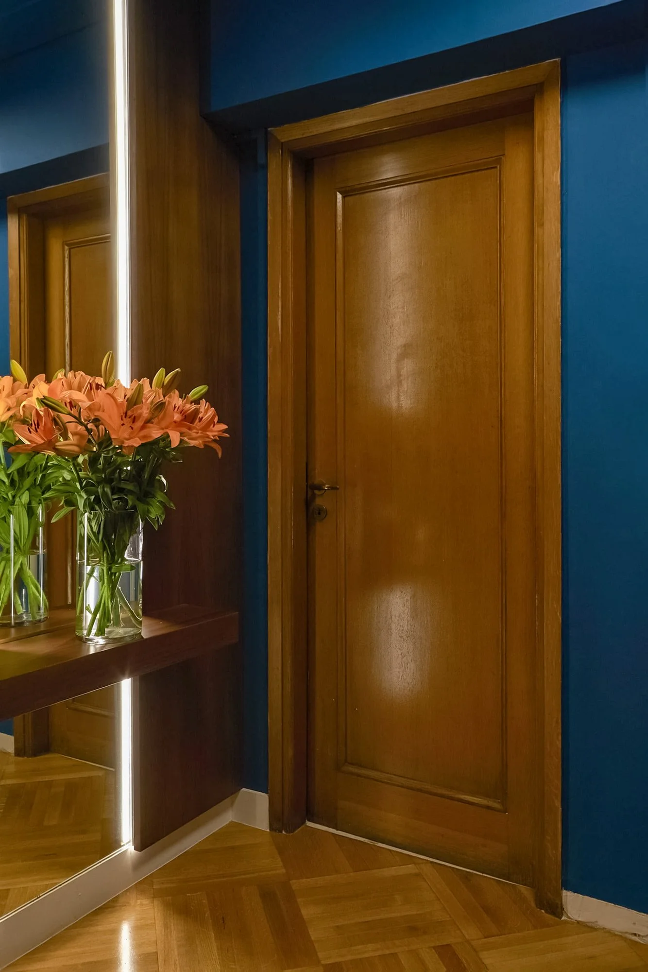 First view of a luxury law firm entrance in Kolonaki. Features a solid wood door, deep teal accent walls, and a minimalist lit mirror with fresh orange lilies. Interior design by Sissy Feida, photography by Stavros Kostakis