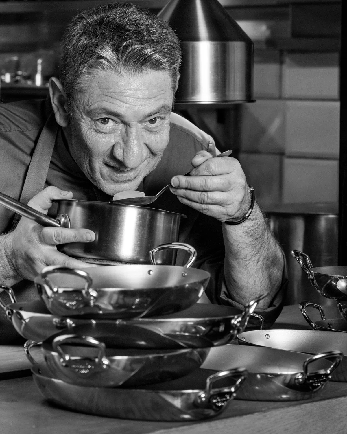 Cinematic black-and-white editorial portrait of Chef Alain Parodi in his busy kitchen, focused on tasting from a saucepan with a spoon, surrounded by stacked copper pots, smoke.Professional portrait photography by Stavros Kostakis
