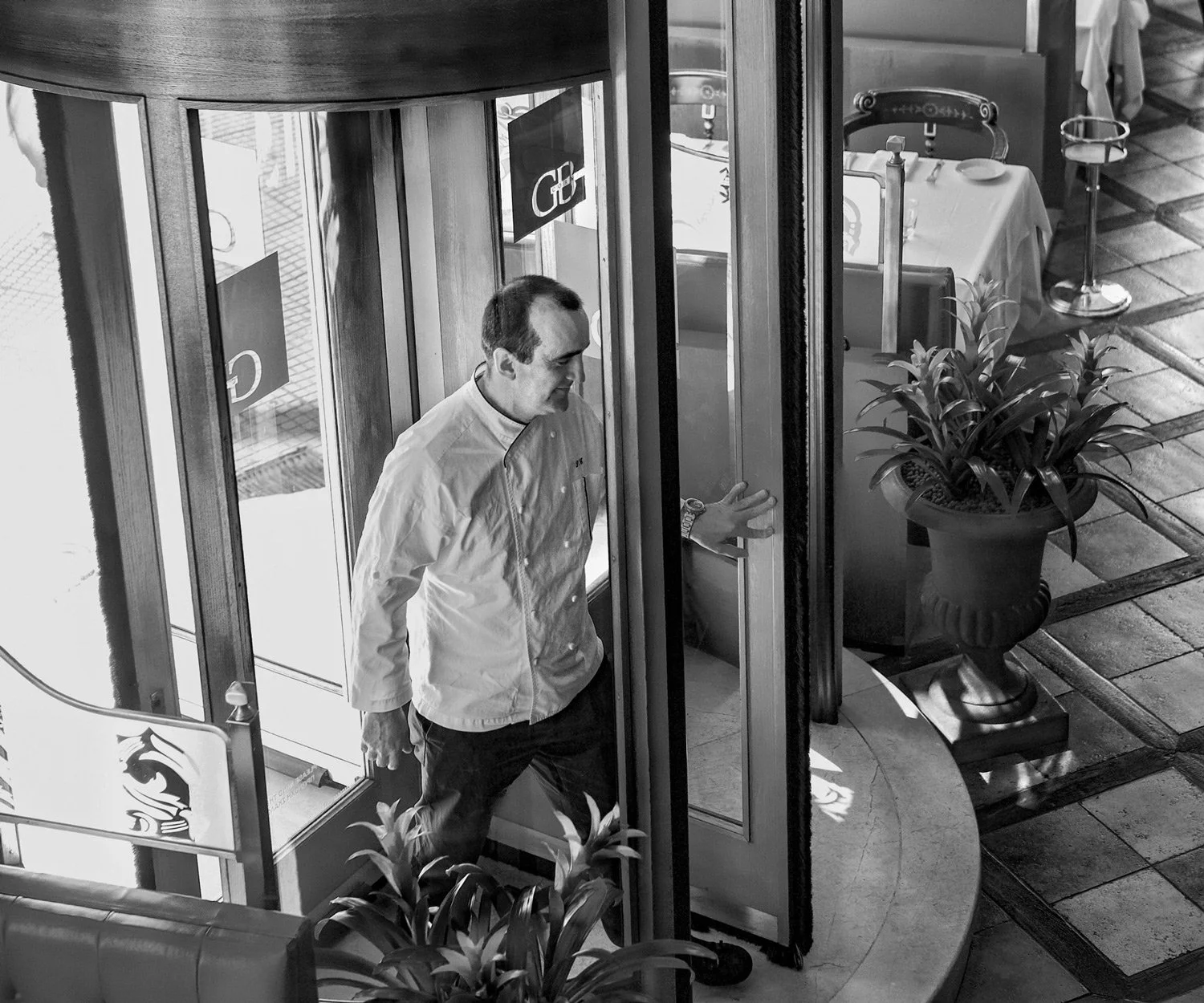 The relentless energy of high-end gastronomy. This dynamic, high-angle black and white portrait of Sotiris Evagelou in motion inside the iconic revolving doors of Grande Bretagne breaks the mold of traditional executive portraits. It's a study in mov