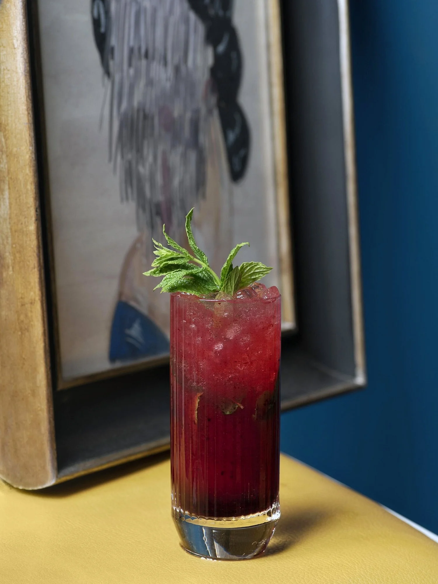 A vibrant red cocktail with fresh mint sprigs in a tall ribbed glass, set against a teal blue wall and a gold-framed artwork at Royal Monkey. Minimalist and atmospheric lifestyle photography by Stavros Kostakis.