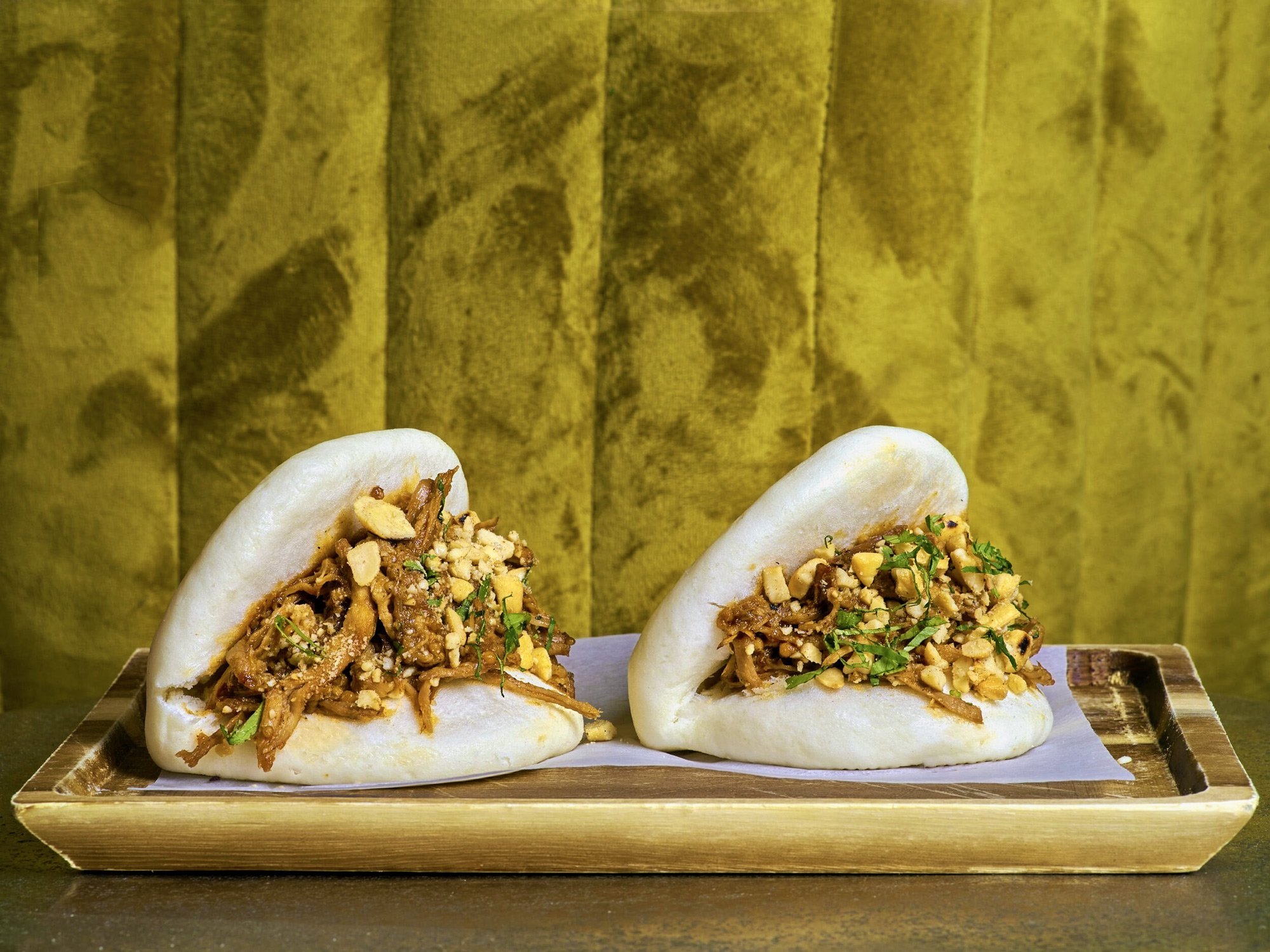 A close-up photograph of two pulled pork Bao Buns garnished with crushed peanuts and fresh coriander, served on a wooden tray. Shot against a textured mustard-yellow velvet background at Royal Monkey Kifisia. Captured by Stavros Kostakis.