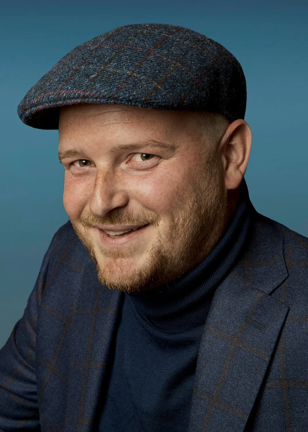 Modern color corporate portrait of Danny Schoorel in Rotterdam, wearing a stylish flat cap. A professional and approachable executive headshot by Stavros Kostakis, focusing on personal branding and creative business identity with a clean blue backgro