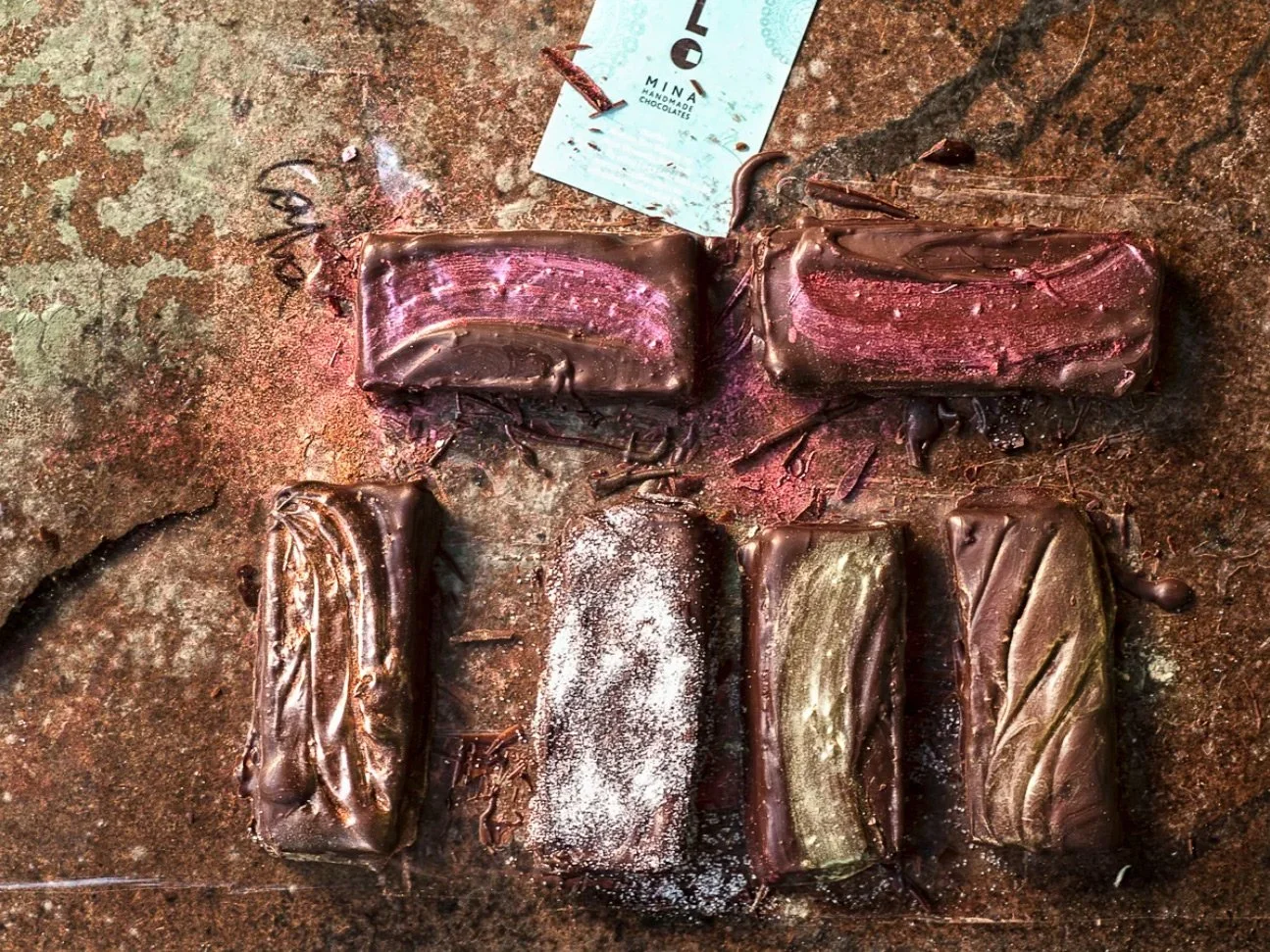 Commercial food photography & styling by Stavros Kostakis for Mina Postolidi featuring handmade chocolates with gold and pink metallic accents and the brand's identity card on a textured surface.