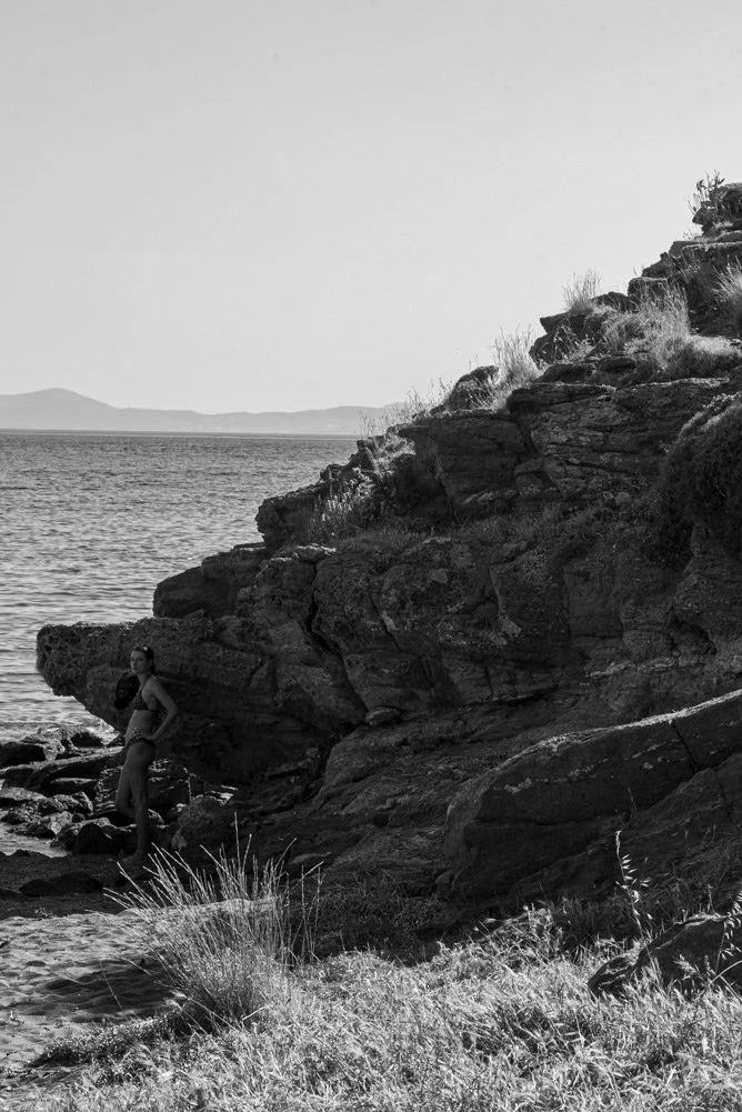 B&W cinematic portrait of a woman by the rugged cliffs of Vourni, Tinos; raw 1960s aesthetic and timeless Aegean nature by Stavros Kostakis