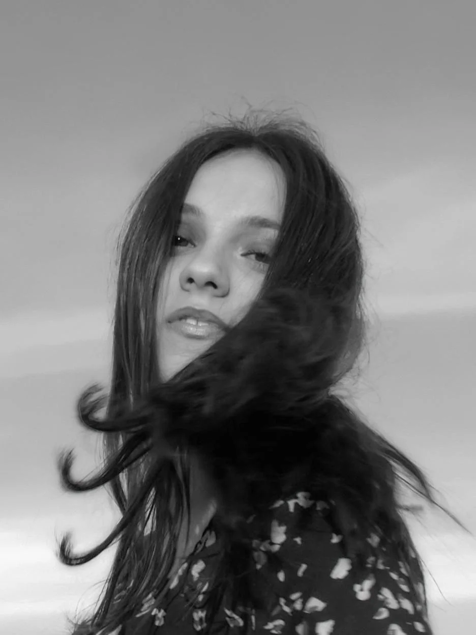 Maria Sanida | Cinematic black and white portrait of a young woman with wind-blown hair, captured in a raw and authentic moment. The photograph emphasizes natural beauty and emotional depth through soft lighting and a documentary aesthetic. A fine ar