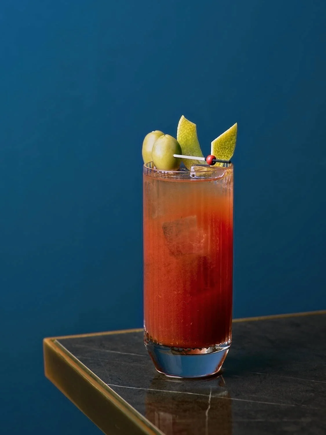 A vibrant red cocktail garnished with green olives on a gold-rimmed table against a teal blue wall at Royal Monkey. Minimalist lifestyle photography by Stavros Kostakis.