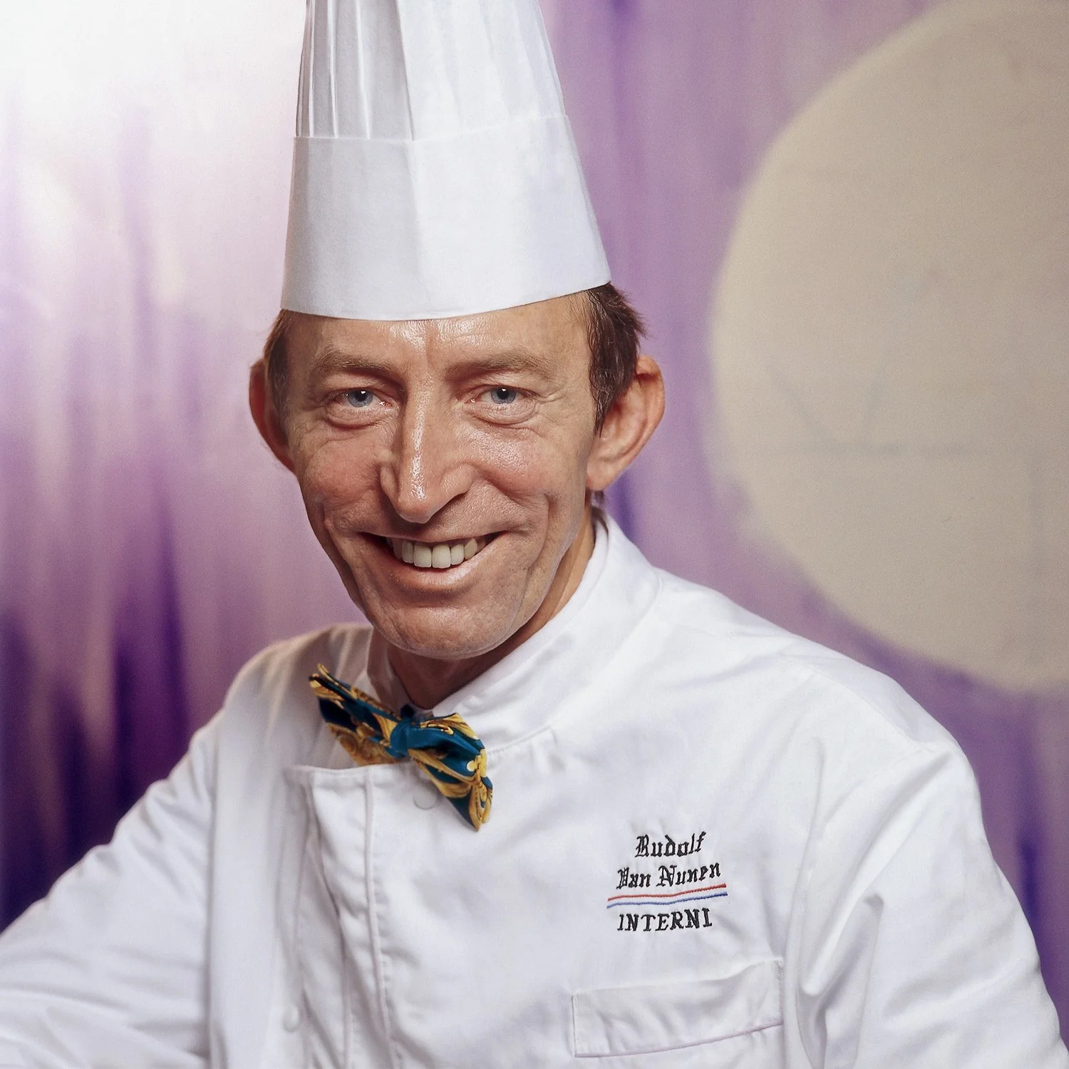 Close-up editorial portrait of smiling Chef Rudolf van Nunen in his professional white jacket and signature bow tie. A warm and authentic character study by Stavros Kostakis, capturing the personality and expertise of a world-class chef.