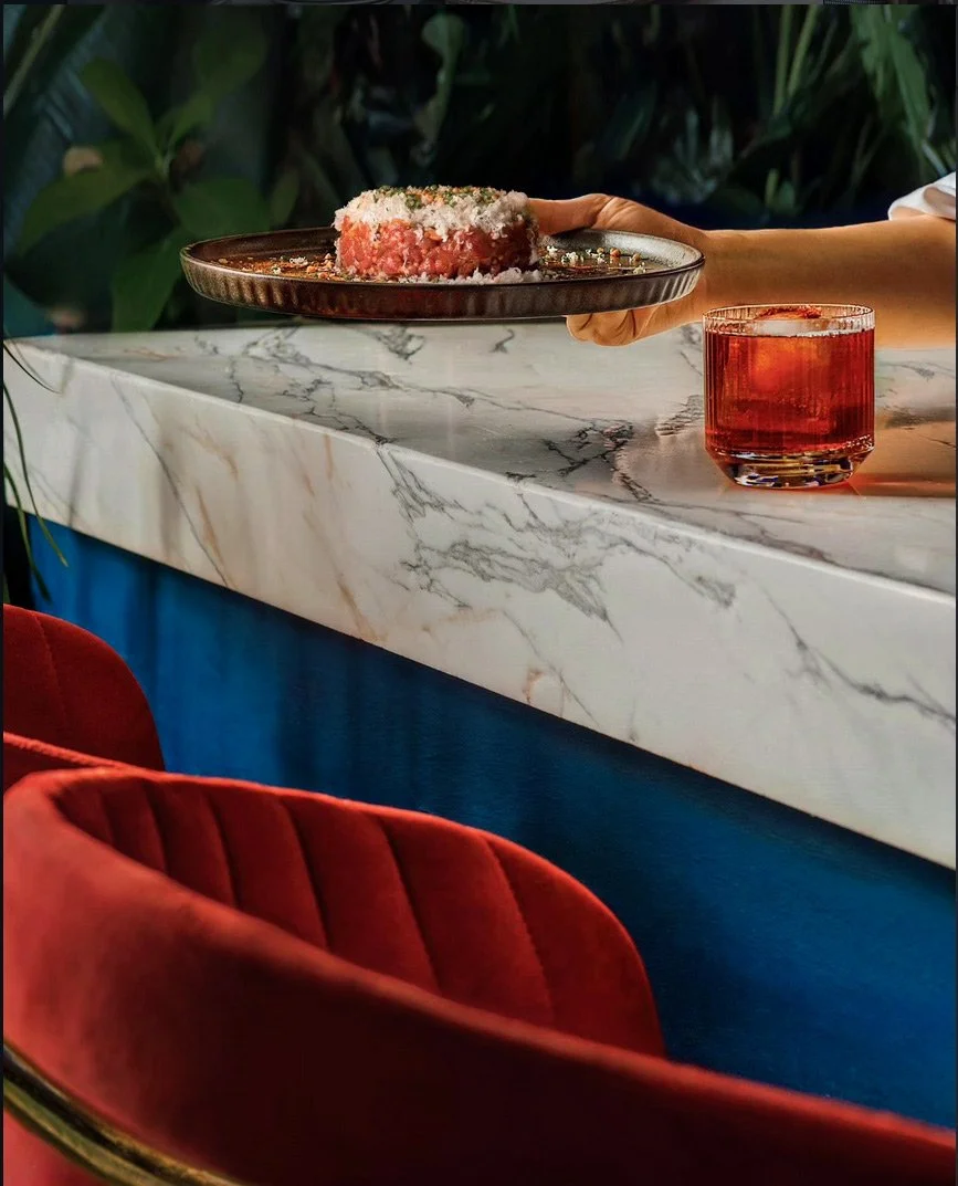 A dynamic lifestyle gastronomy shot showing a gourmet beef tartar dish being served on a textured platter next to a classic Negroni cocktail with a clear ice cube. Captured over a blue bar counter with a vibrant red chair in the foreground at Royal M