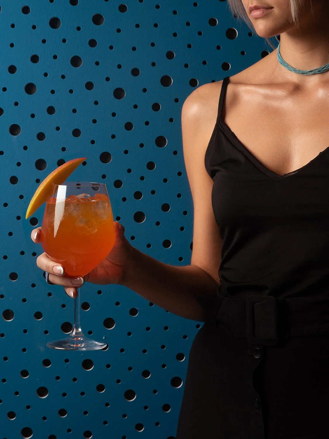 A candid, low-key lighting lifestyle shot at Royal Monkey Kifisia. A young woman's hand is holding a sophisticated amber cocktail with an orange garnish, while sitting next to a gourmet double burger. Atmospheric gastronomy photography capturing a mo