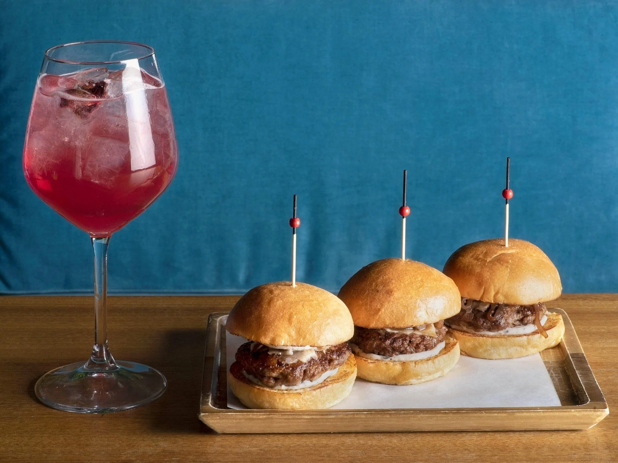 Three mini gourmet burger sliders on a wooden tray accompanied by a refreshing red spritz cocktail. Shot by Stavros Kostakis for Royal Monkey's brand assets.