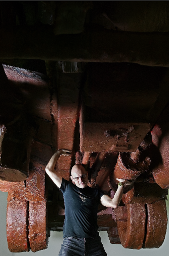Greek contemporary artist Poka-Yio standing beneath his massive chocolate Steyr vehicle sculpture. Dramatic low-angle portrait capturing the scale of the art installation. Photography by Stavros Kostakis.