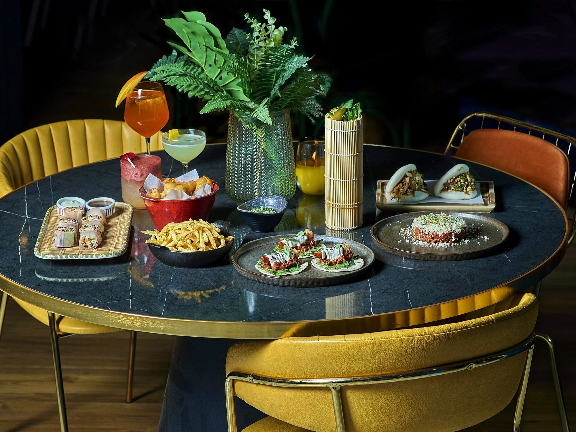 A grand wide shot of a round black marble table at Royal Monkey Kifisia, filled with a fusion feast including beef tartar, tacos, spring rolls, and signature cocktails, surrounded by mustard yellow velvet chairs. Visual storytelling by Stavros Kostak