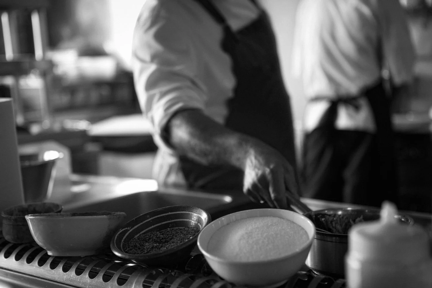A cinematic black and white documentary photograph by Stavros Kostakis, showing the focused atmosphere and raw ingredient prep inside a professional kitchen.