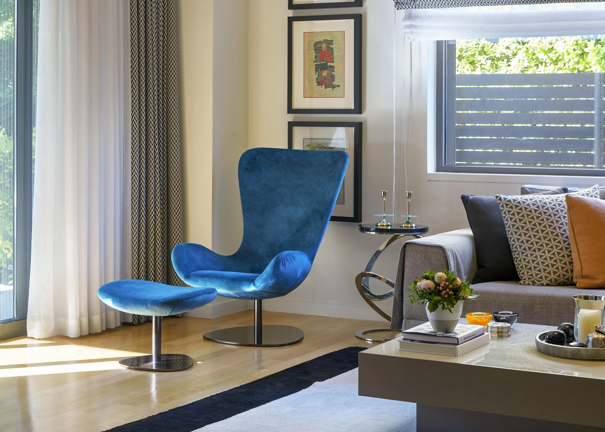 A stylish corner of a contemporary Ekali living space. Anchored by a mid-century modern inspired blue velvet lounge chair and matching ottoman on a graphic geometric rug. Features layered textures, graphic curtains, curated wall art, and elegant coff
