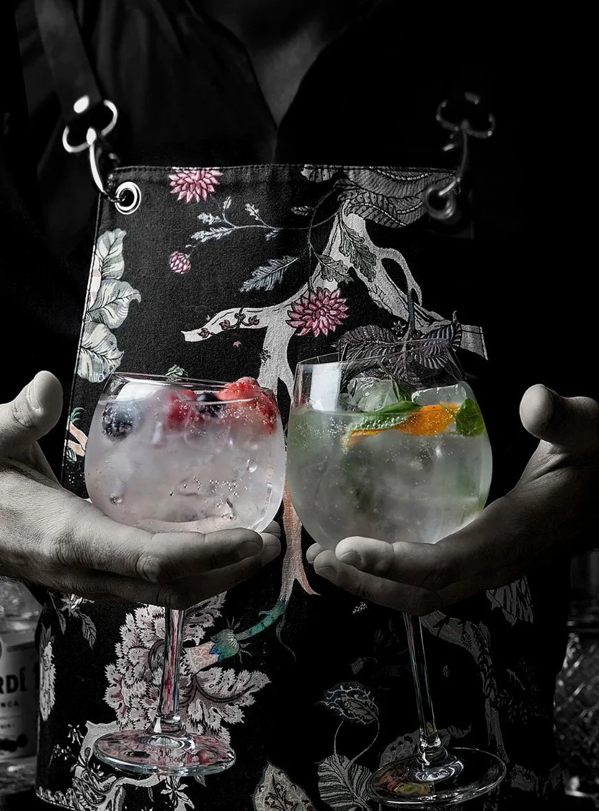 Irodion Capelle | Rotterdam Culinary dining Irodion Capelle | High-end mixology photography for Irodion Capelle. Close-up of a bartender holding two gourmet gin cocktails with fresh berries and herbs, wearing a decorative floral apron. Dramatic dark 