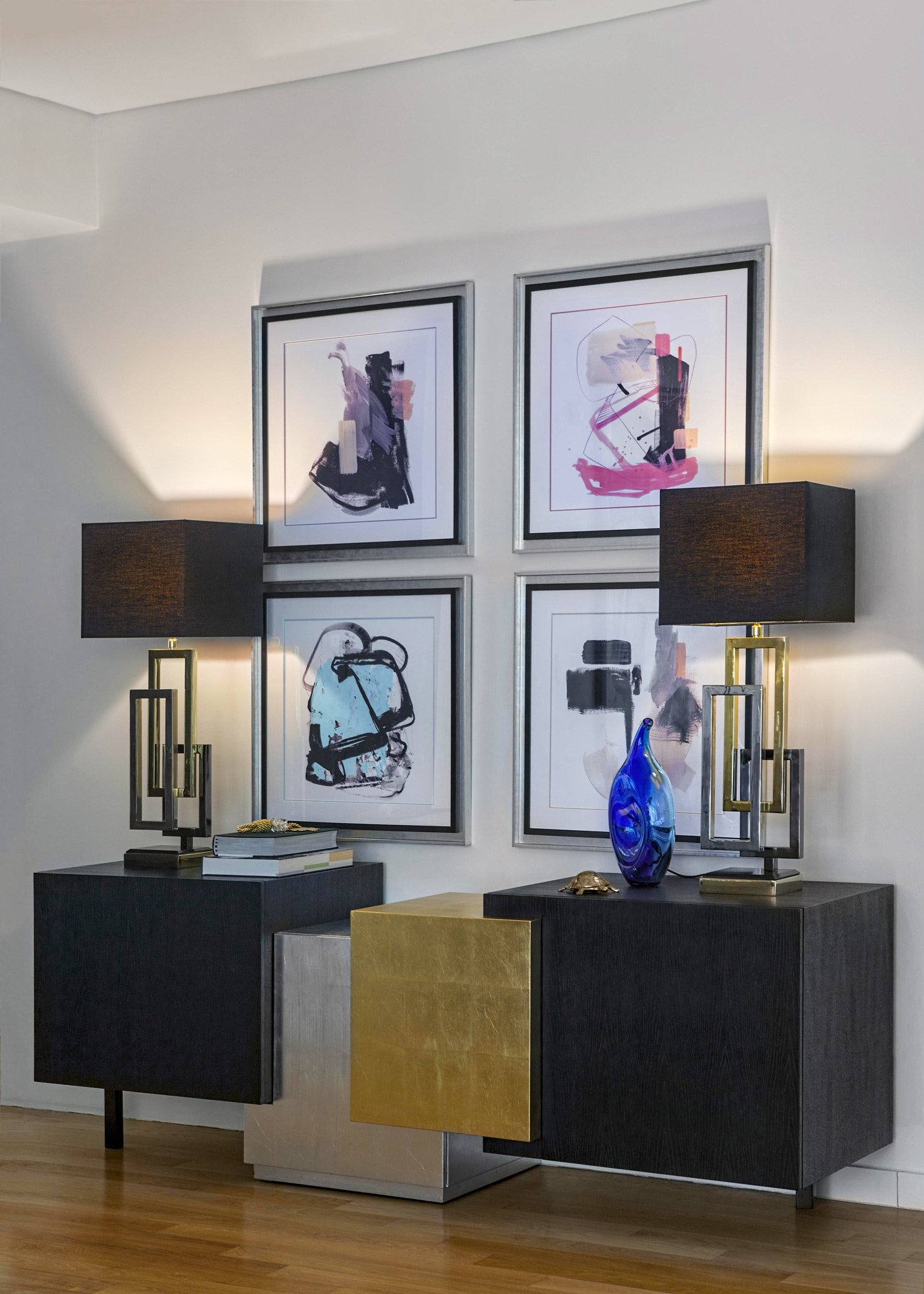 A perfectly symmetrical high-end interior vignette from an Ekali residence. Features two bespoke black and gold sculptural table lamps with ornate shades, flanking a curated 2x2 grid of abstract wall art. A dark textured console and a striking blue c