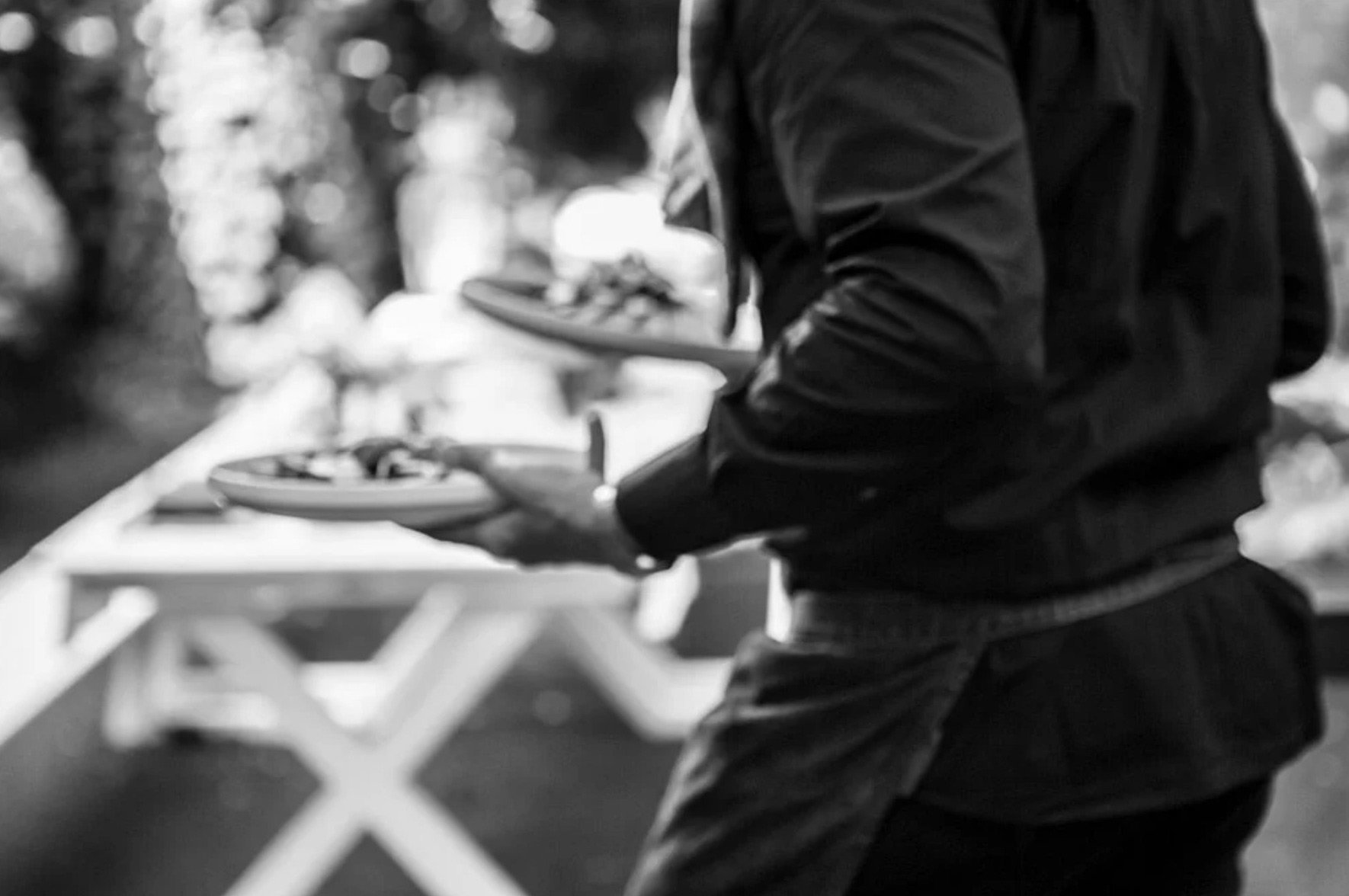 A black and white documentary-style photograph by Stavros Kostakis showing a waiter in motion carrying plates during a high-end dining event in Brussels.