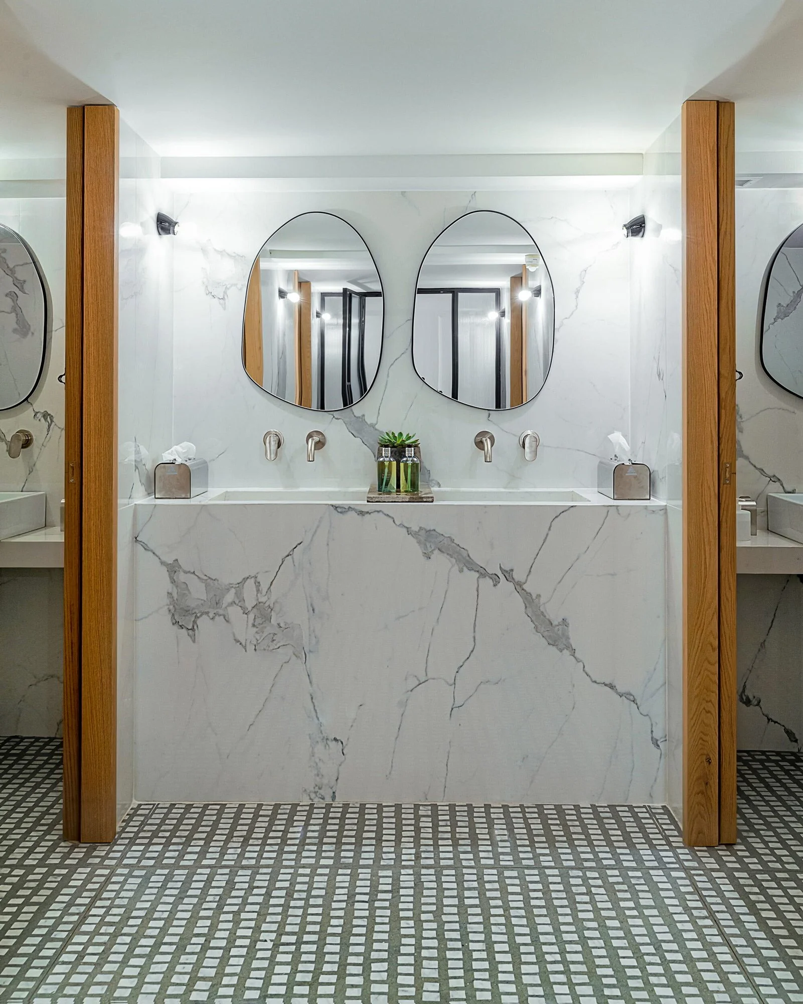 A luxury restaurant restroom designed by Papaioannou, featuring floor-to-ceiling white marble with grey veining, organic-shaped mirrors, and warm wood architectural pillars. The mosaic floor adds a rhythmic texture to the minimalist and sophisticated